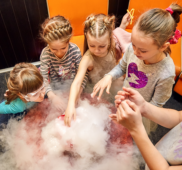 Group of kids reaching out to touch science experiment on table giving off vapor