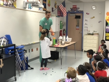 Young girl in eye goggles and white lab coat volunteering during science show