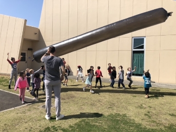 Doctor Larson holding a long dark plastic object outdoors with a class of kids